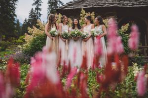 Bridesmaids in blush gowns holding bouquets stand together in a lush garden with pink flowers in the foreground.