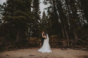 Bride and groom share a romantic embrace surrounded by tall pine trees on a sandy forest path.