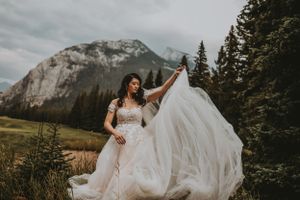 Bride in a lace gown tosses her veil dramatically in a mountain meadow with rocky peaks behind her.