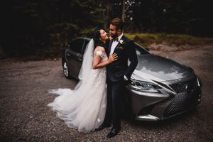 Bride and groom pose elegantly in front of a silver Lexus luxury car in a forest setting.
