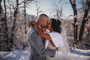 Bride and groom share a passionate kiss surrounded by snow-covered trees in a winter wonderland.