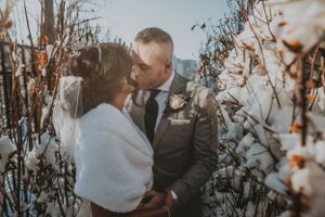 Bride in a white fur wrap and groom kiss tenderly among cotton plants dusted with winter snow.
