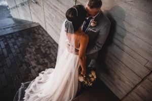 Bride and groom share an intimate moment leaning against a concrete wall, bouquet in hand.