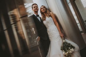 Elegant bride in lace mermaid gown and groom in tuxedo pose together near a window with bouquet.