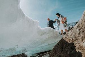 Adventurous wedding couple climbing snowy rocky terrain under bright blue sky during mountain elopement.