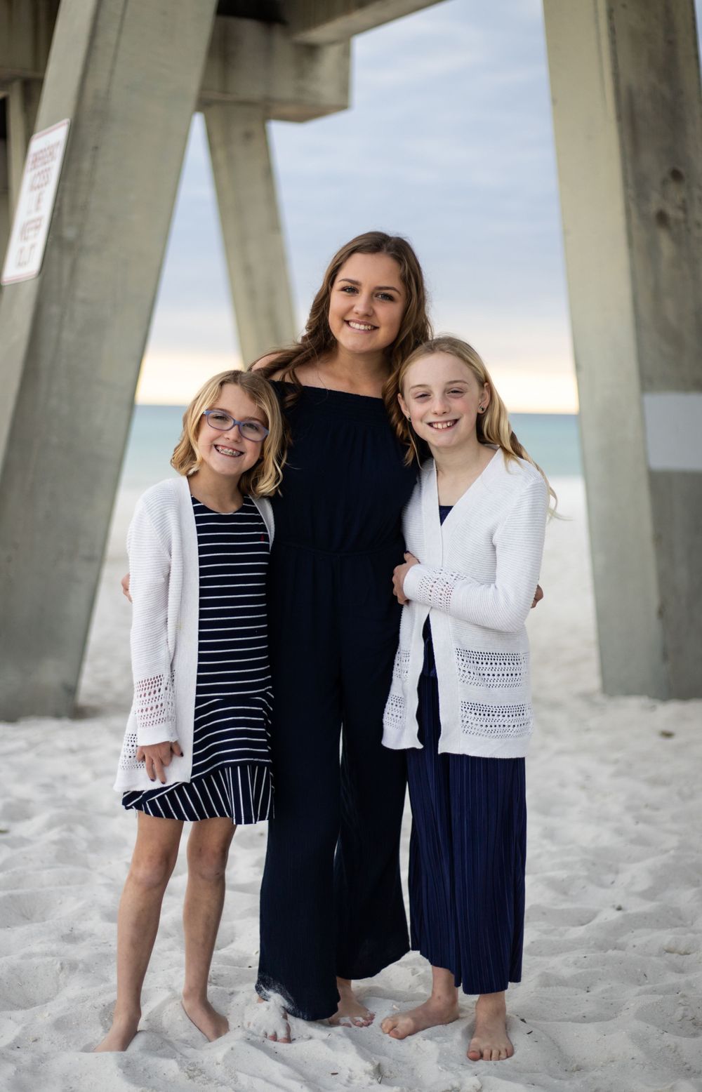 Three sisters posing together under beach pier on sandy shore, wearing coordinated navy and white outfits.