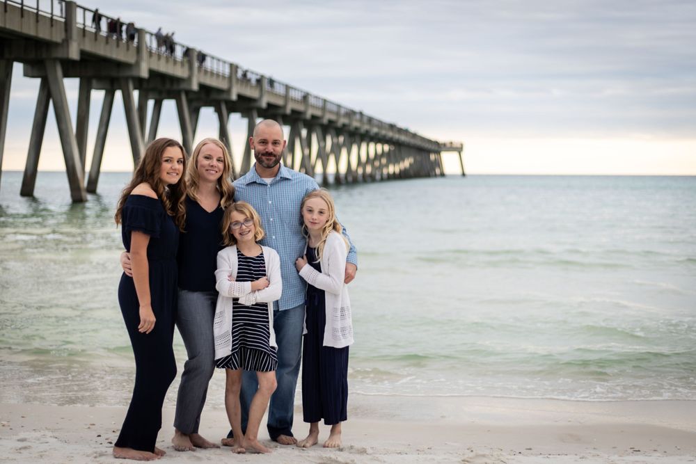 Family of five smiling together on beach near pier, parents with three daughters on sandy shore at dusk.