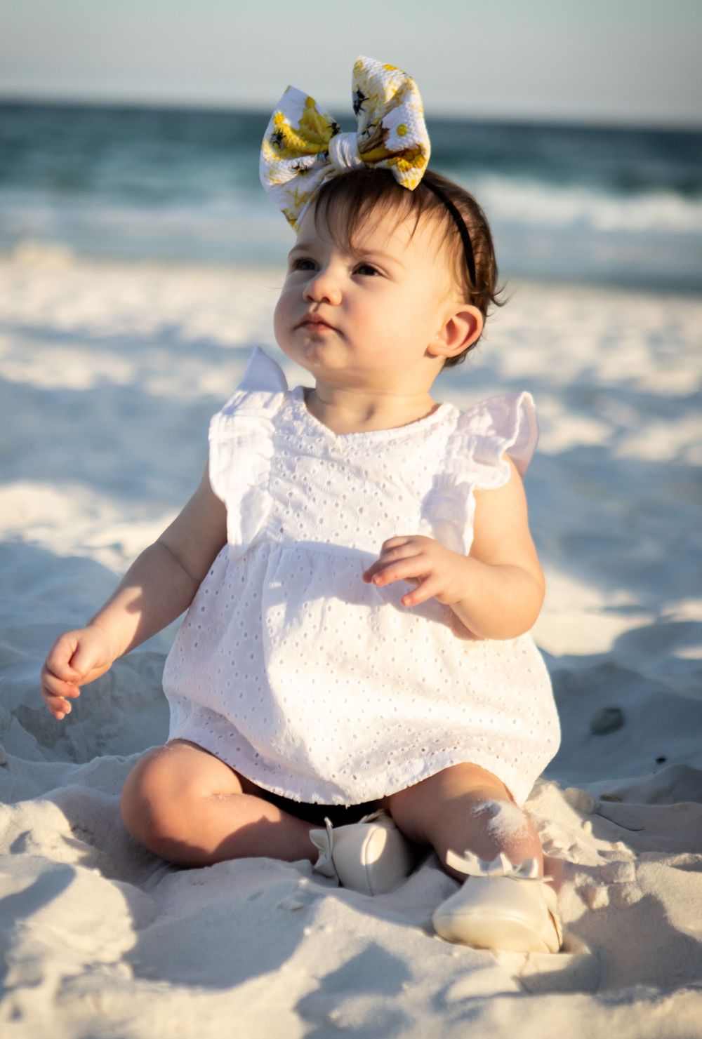 Adorable baby girl in white dress with gold bow sitting on beach sand.