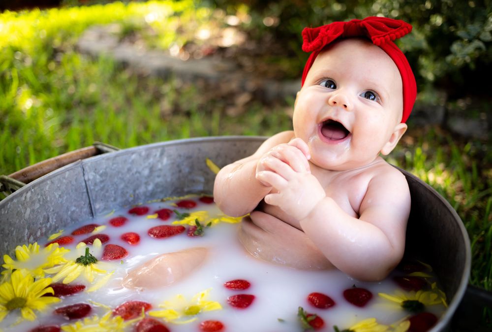 Cute baby girl with red bow headband laughing in outdoor milk bath with strawberries and yellow flowers in tub.