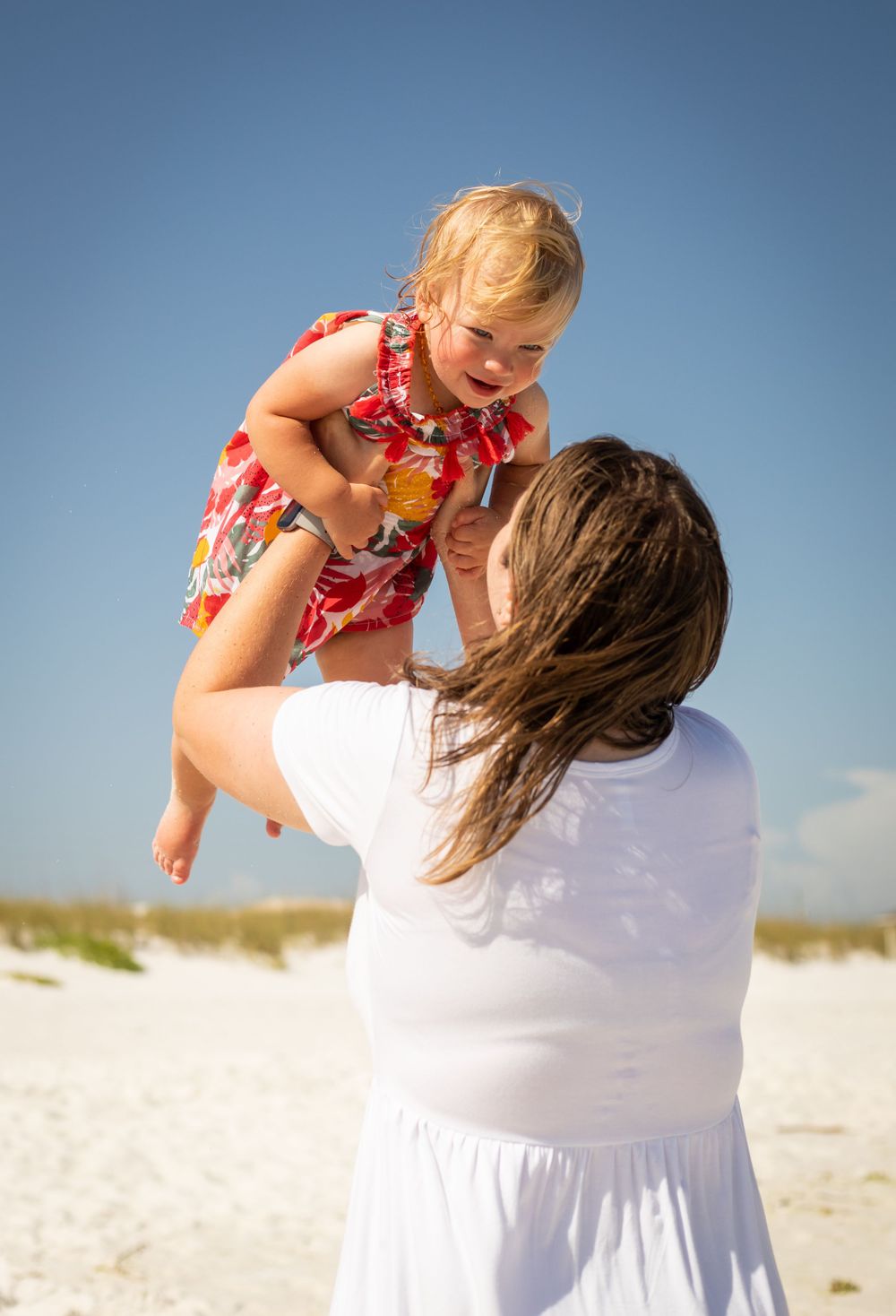 Mother lifting smiling toddler girl in floral dress on a sunny beach.