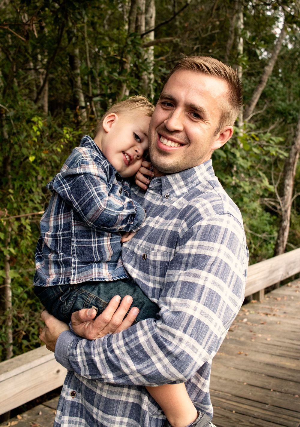 Smiling father holding his young blonde toddler son on a wooden bridge in the forest.