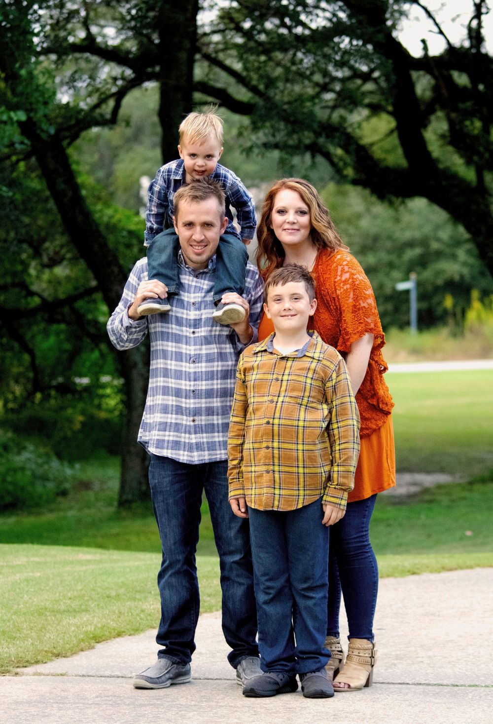 Happy family of four posing together on a park path surrounded by lush green trees.