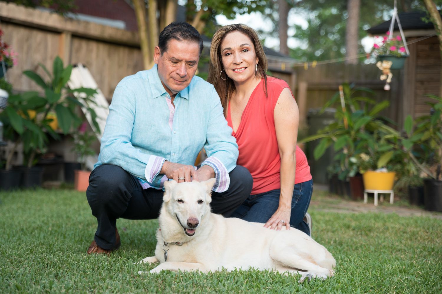 A white dog lies on grass in a backyard garden setting with two people in casual attire.