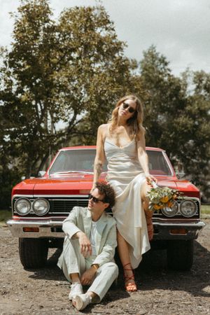 Stylish wedding couple poses with vintage red car in outdoor photoshoot.