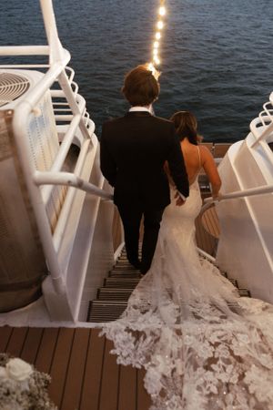 A couple descends white stairs near water at dusk with a flowing wedding dress trailing behind.