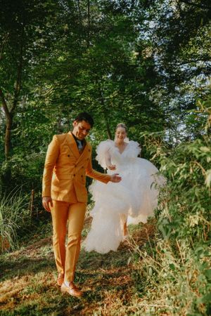 A couple walks through lush greenery wearing white and mustard yellow formal attire.