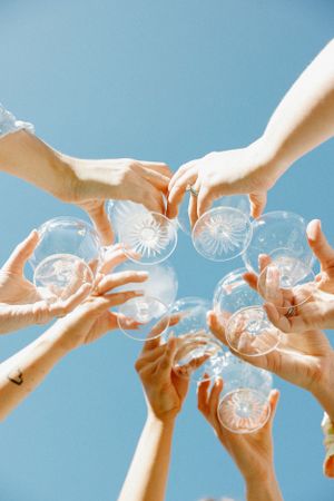 Hands raise champagne glasses against a bright blue sky in a celebratory toast.