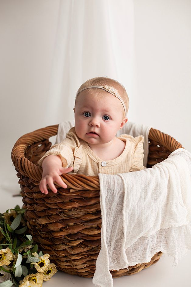 Six month old baby girl sitting in a woven basket with flowers at the bottom.