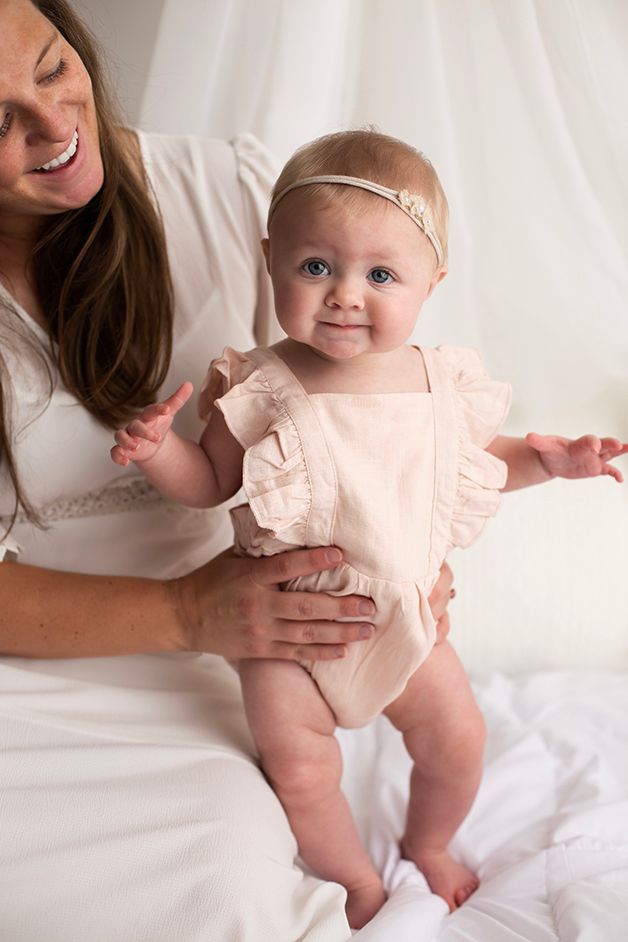 Six month old baby girl sitting on mom's lap.  She is wearing a pink romper and headband.