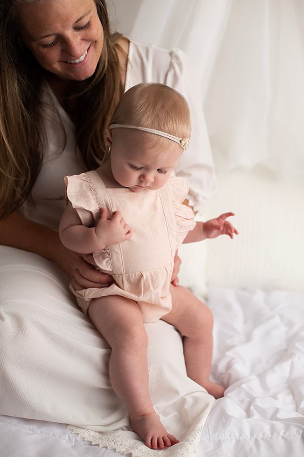 Six month old baby girl sitting on mom's lap wearing a pink romper looking down at her toes.