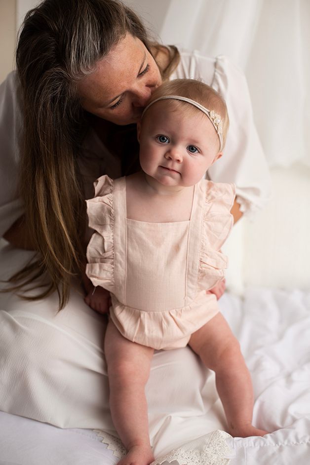 Six month old baby girl sitting on mom's lap in a pink romper.  Mom is kissing baby's head.