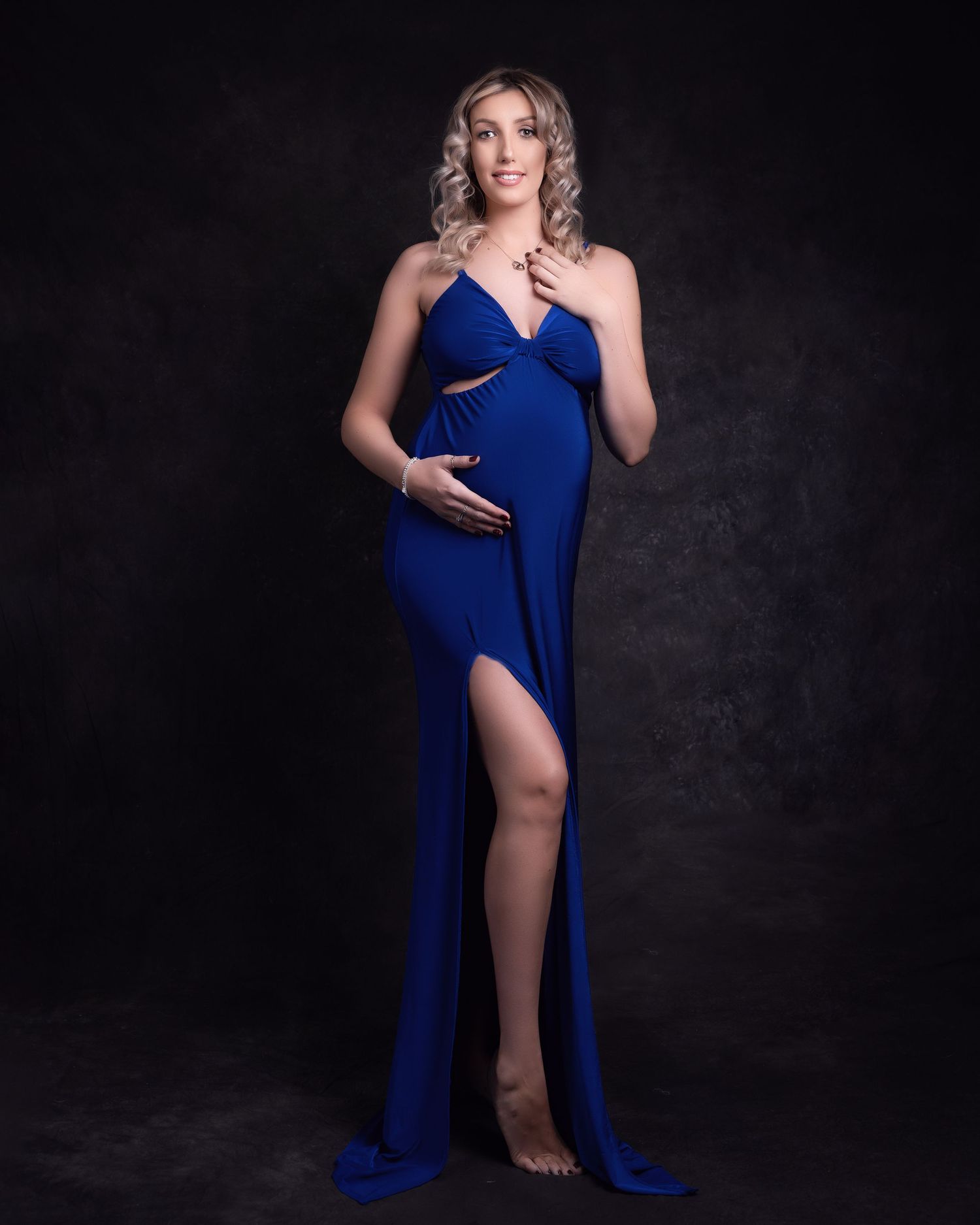 Elegant maternity portrait in a flowing royal blue dress with thigh slit against dark background.