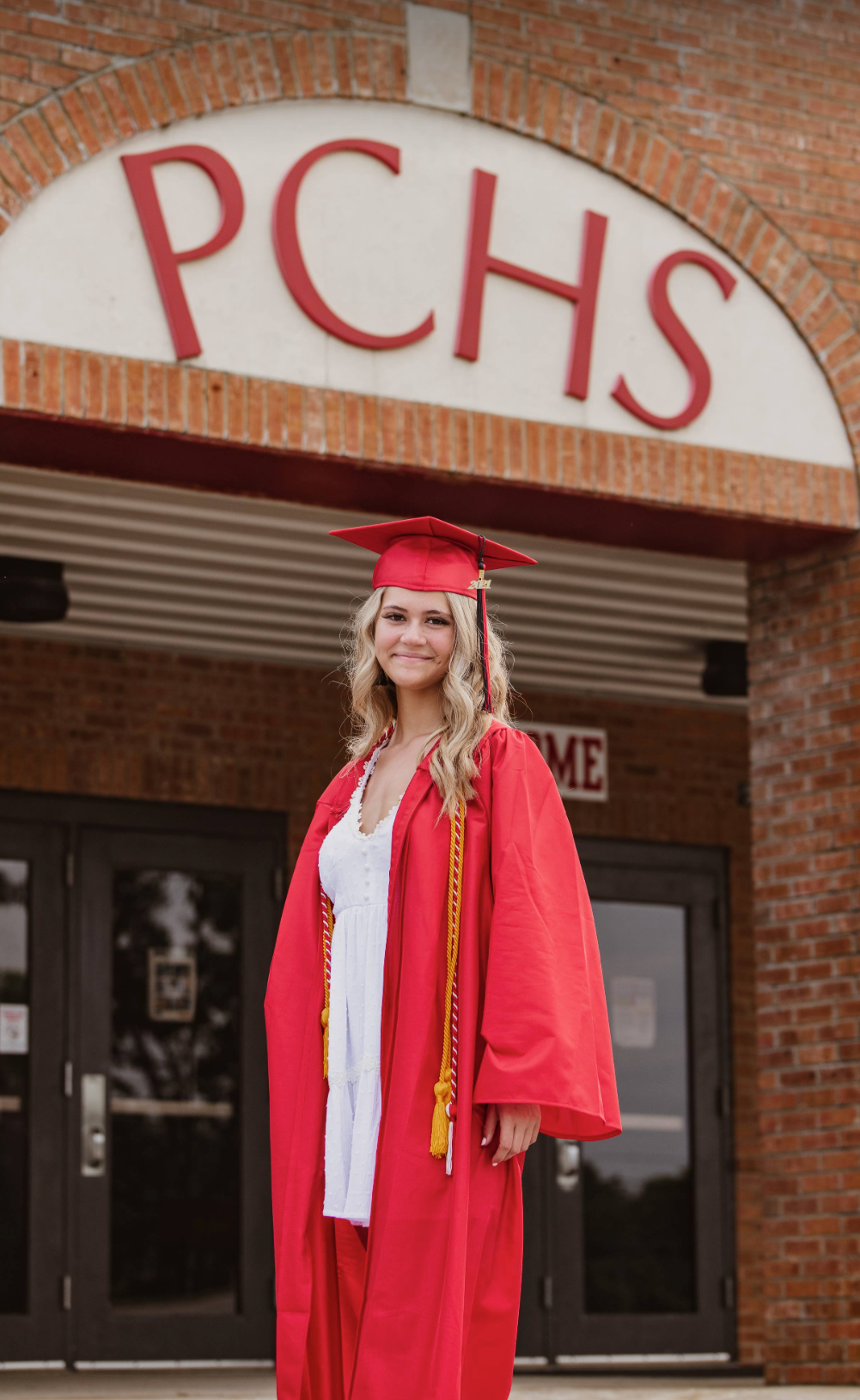 Graduate in red cap and gown poses in front of PCHS brick building entrance.