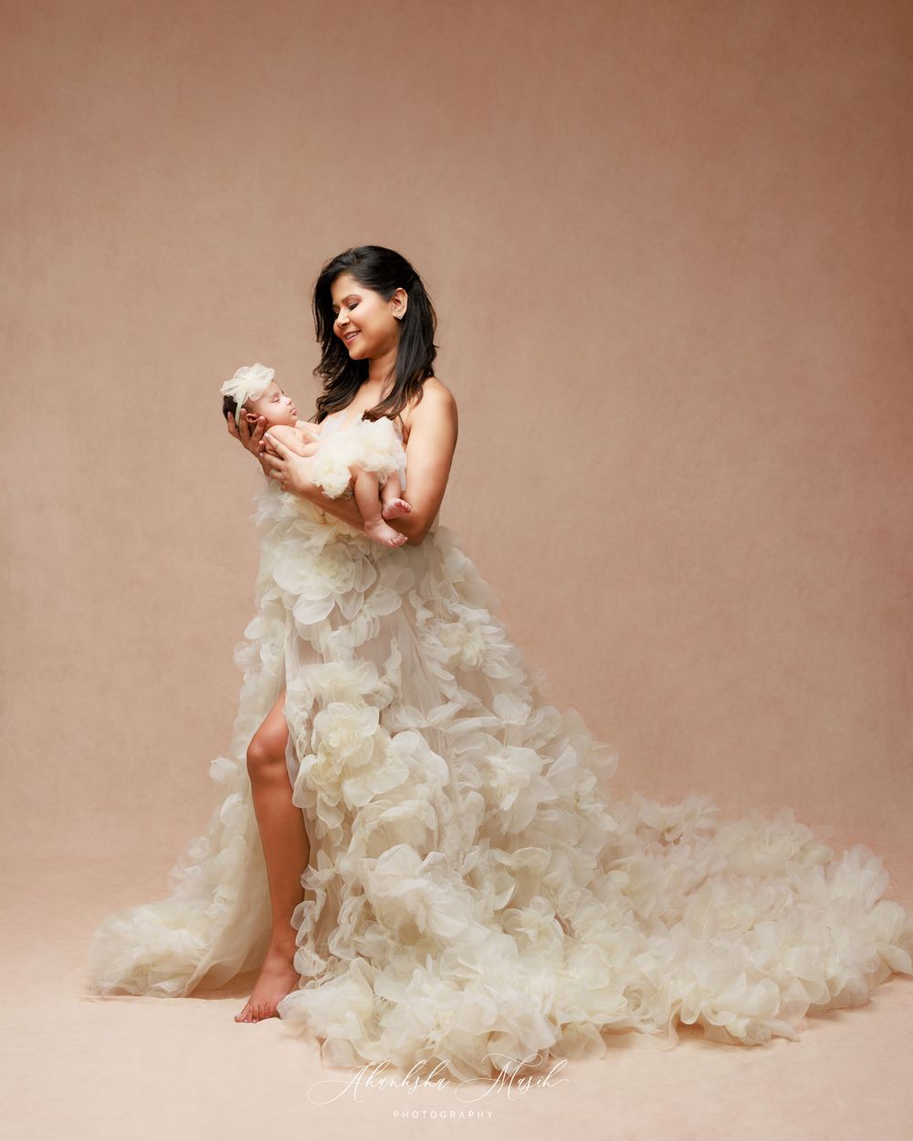 Woman in flowing white ruffled gown holds baby against beige background.