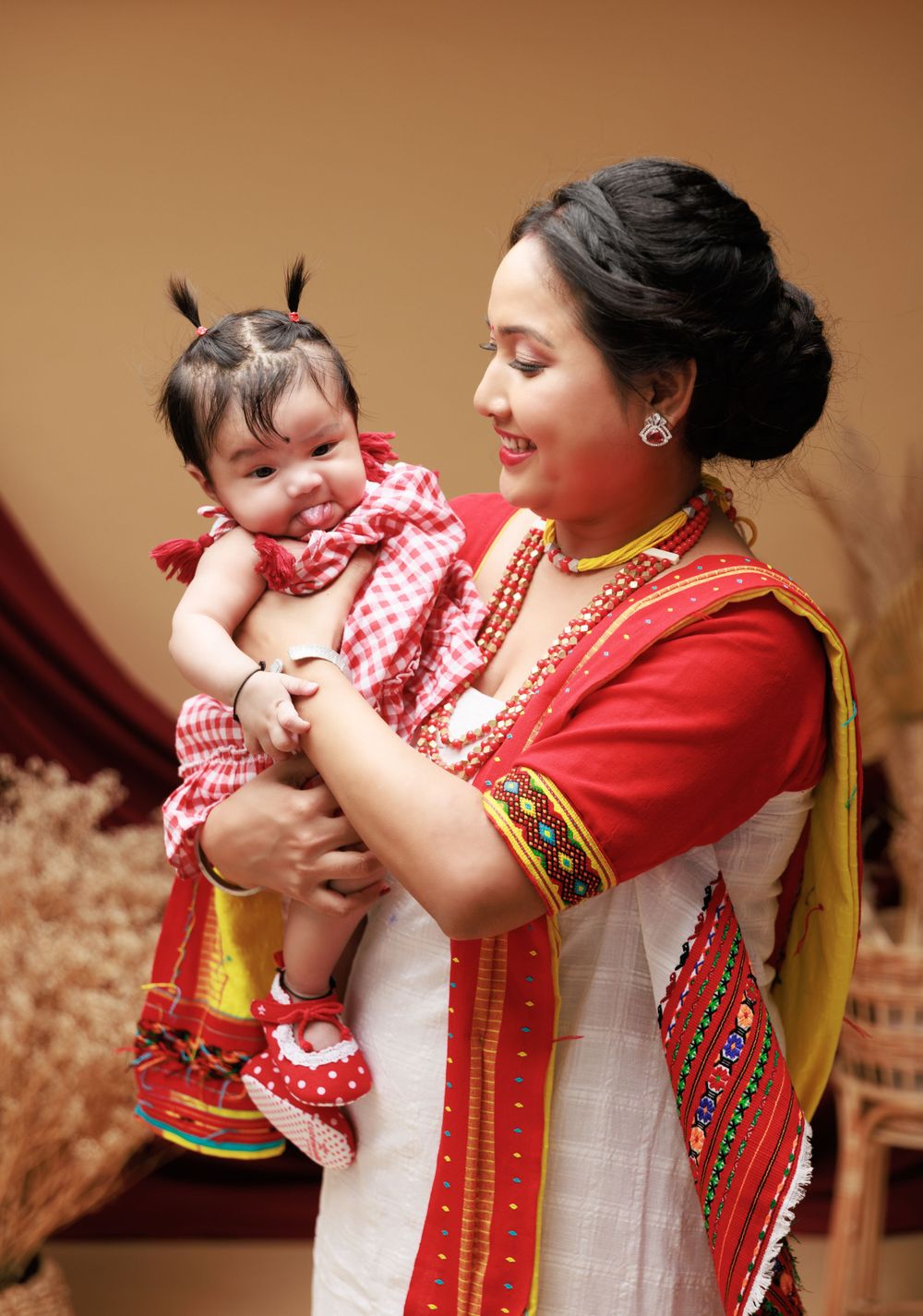 Woman in traditional red attire holds smiling baby in checkered dress.