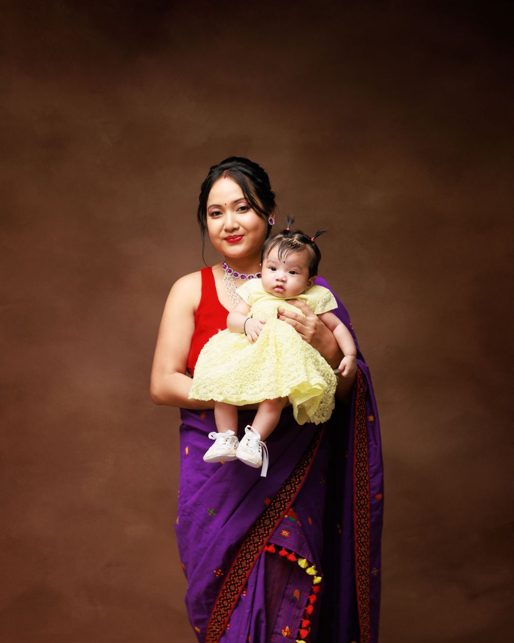 Woman in purple and red sari holds infant wearing yellow dress against brown backdrop.