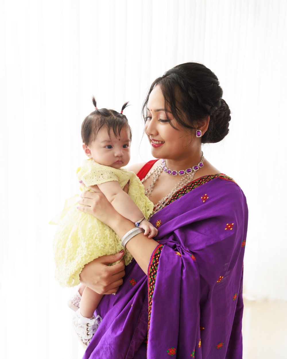 Woman in purple sari holds baby wearing yellow dress against white background.