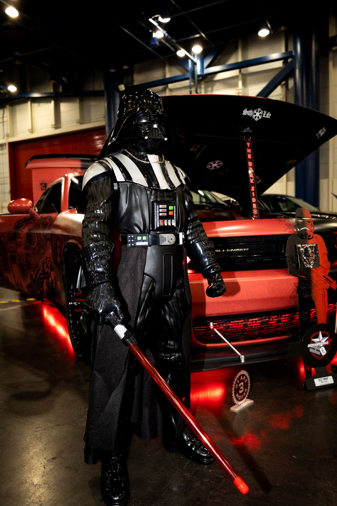 Darth Vader cosplayer with red lightsaber poses next to custom car with red underglow lighting.