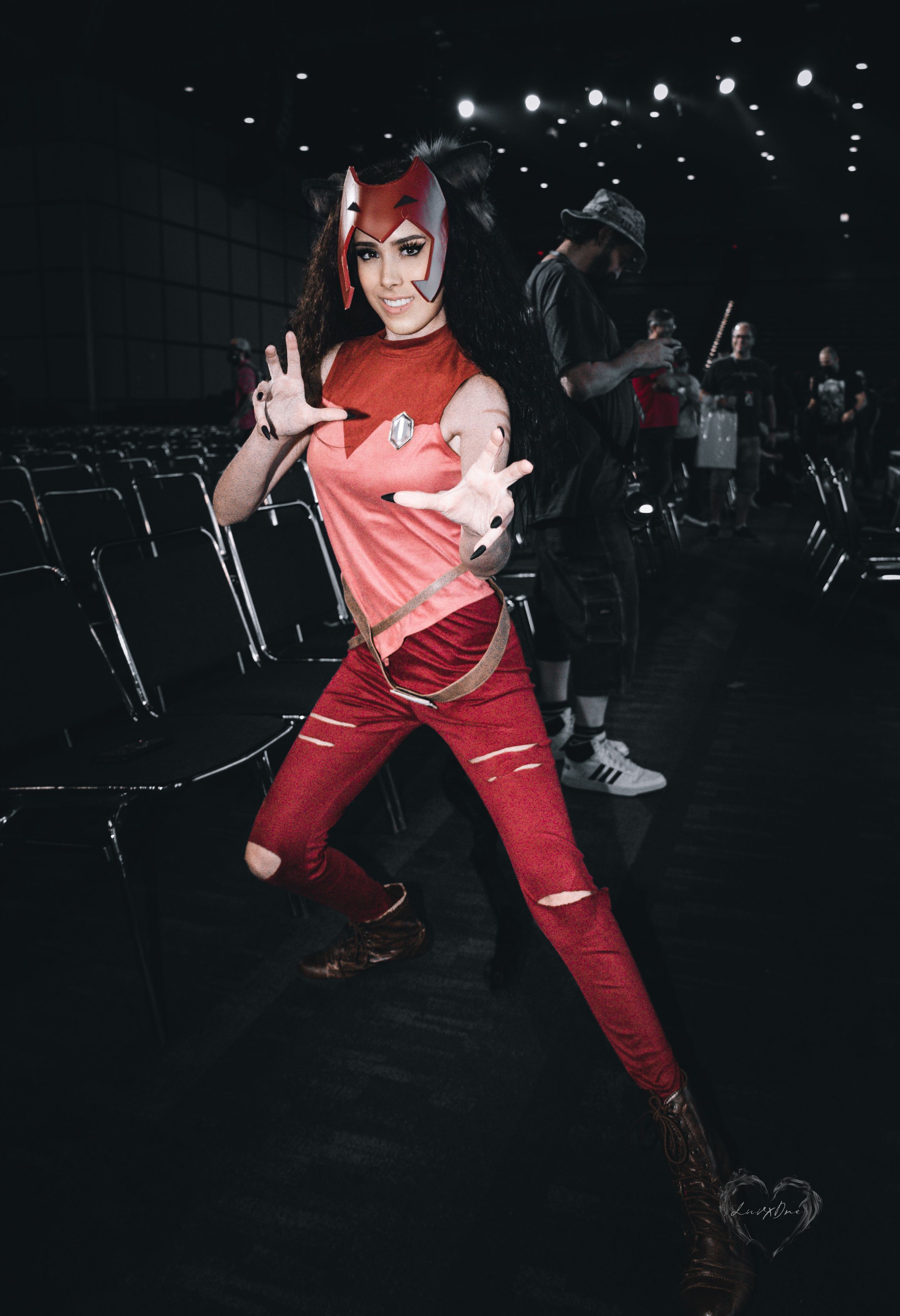 Cosplayer in red outfit strikes action pose in convention center hall with rows of empty chairs visible.