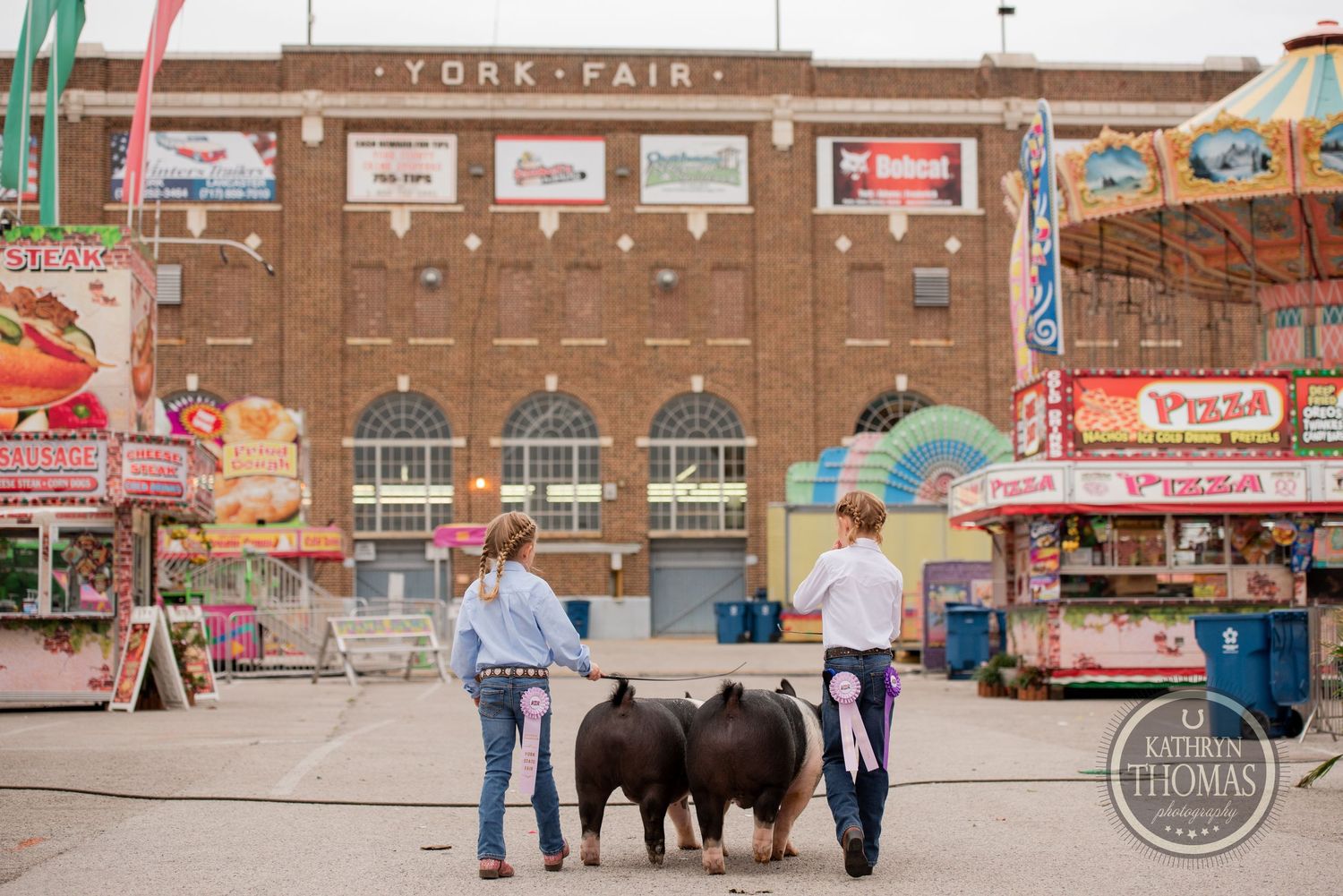 Handlers with livestock stand in front of the York Fair building with carnival rides and food stands.