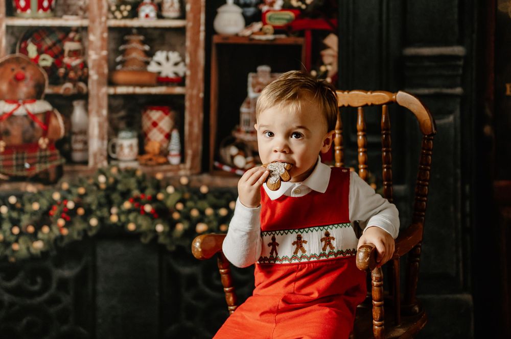 Child in red holiday outfit sitting in wooden chair with Christmas decorations in background.