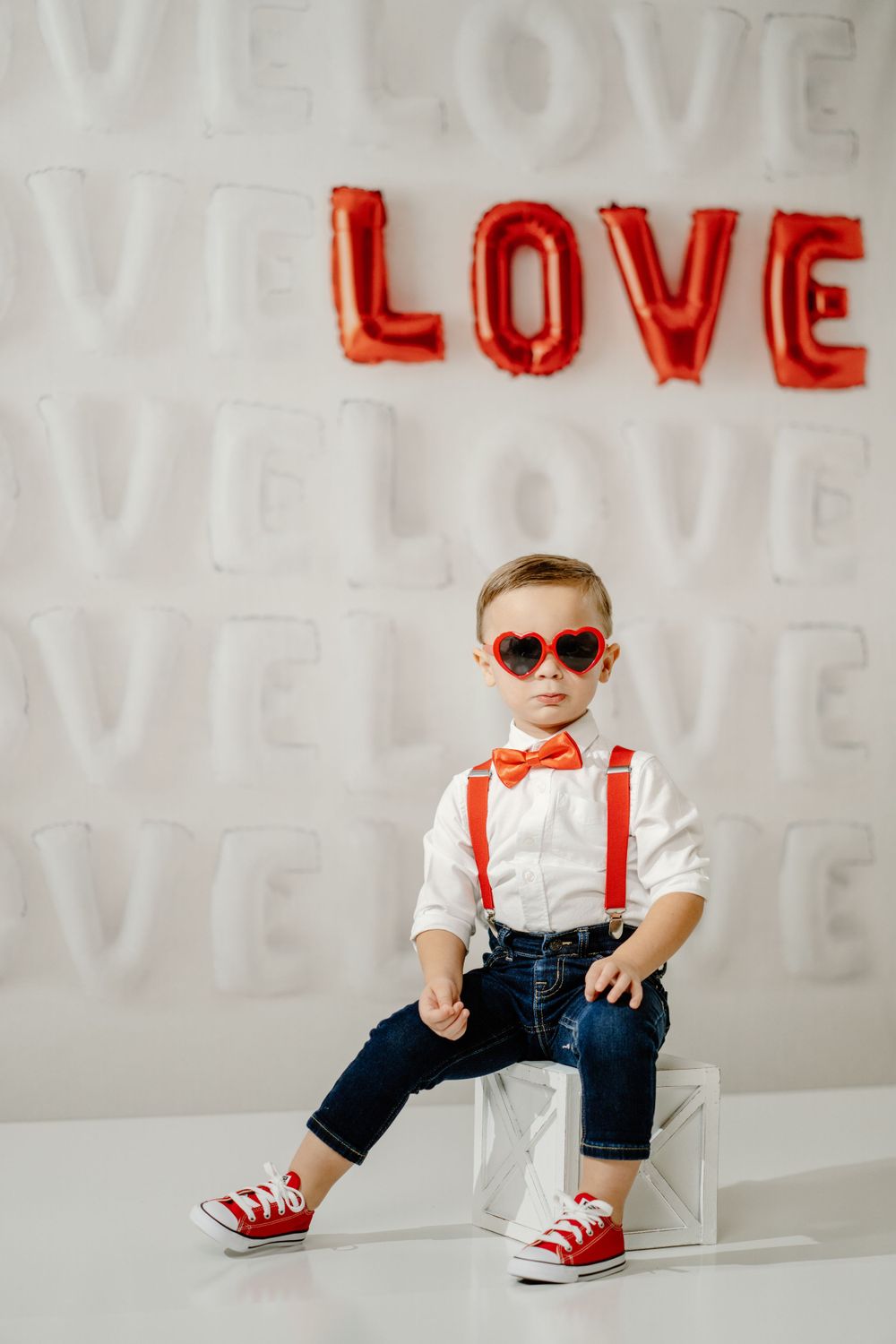 Stylish young boy in sunglasses, suspenders and bow tie poses against white wall with red LOVE balloon letters.