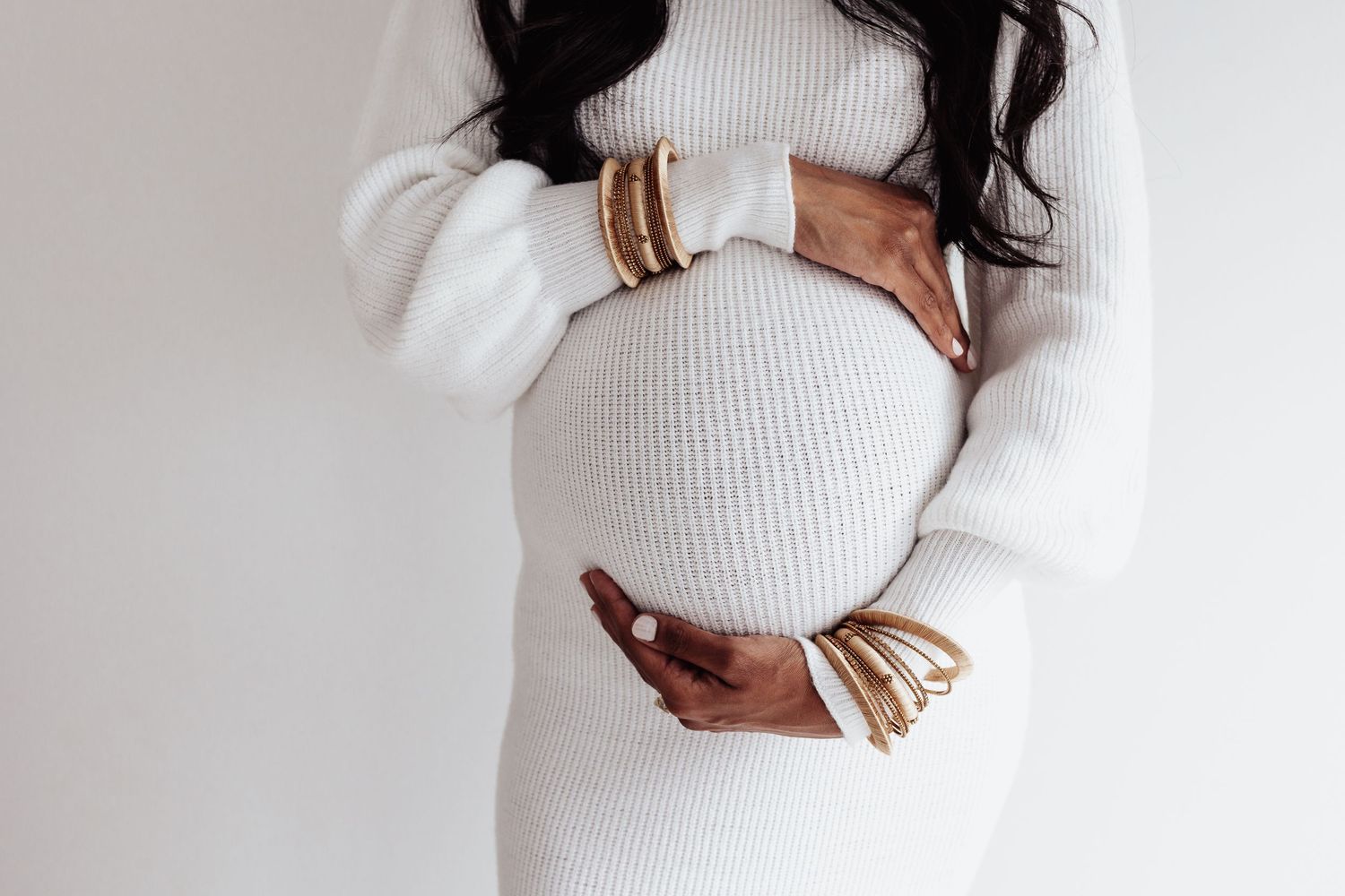 Pregnant woman in white sweater with gold bracelets holding her belly.