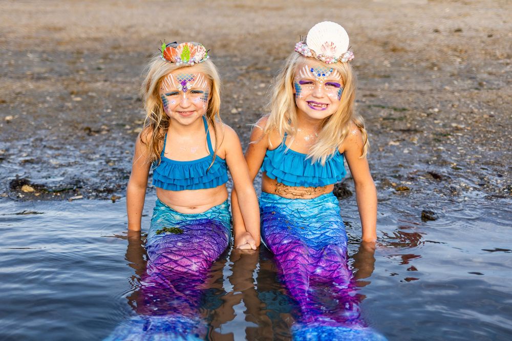 Two mermaids in matching blue and purple costumes pose together in shallow water.