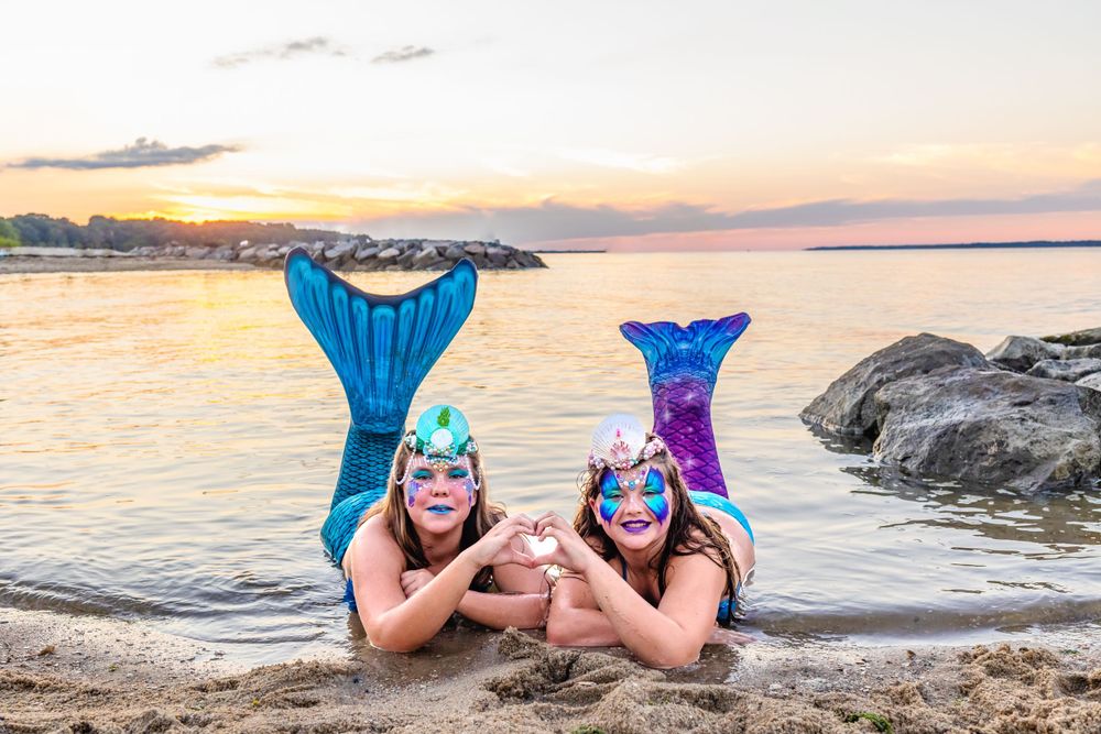 Two mermaids in colorful tails pose on beach during golden sunset.