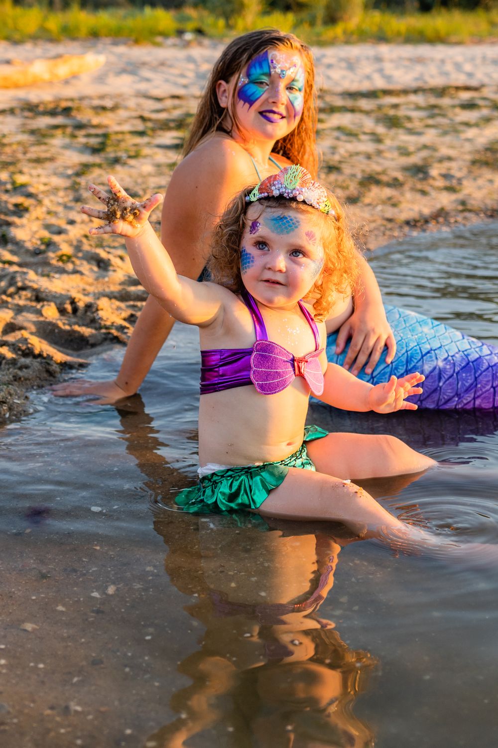 Mermaids in colorful costumes play in shallow water at sunset.