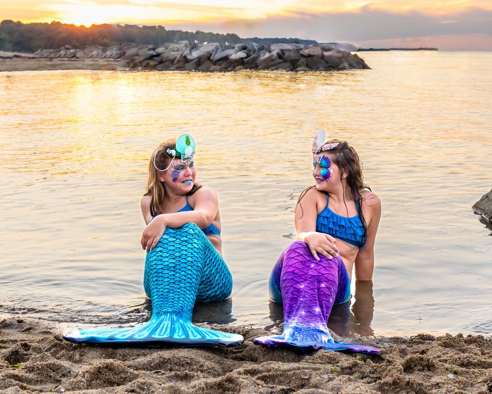 Two mermaids in blue and purple tails rest on beach during sunset.