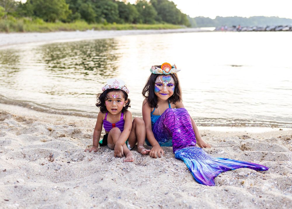 Costumed mermaids in purple tails pose on sandy beach near water.
