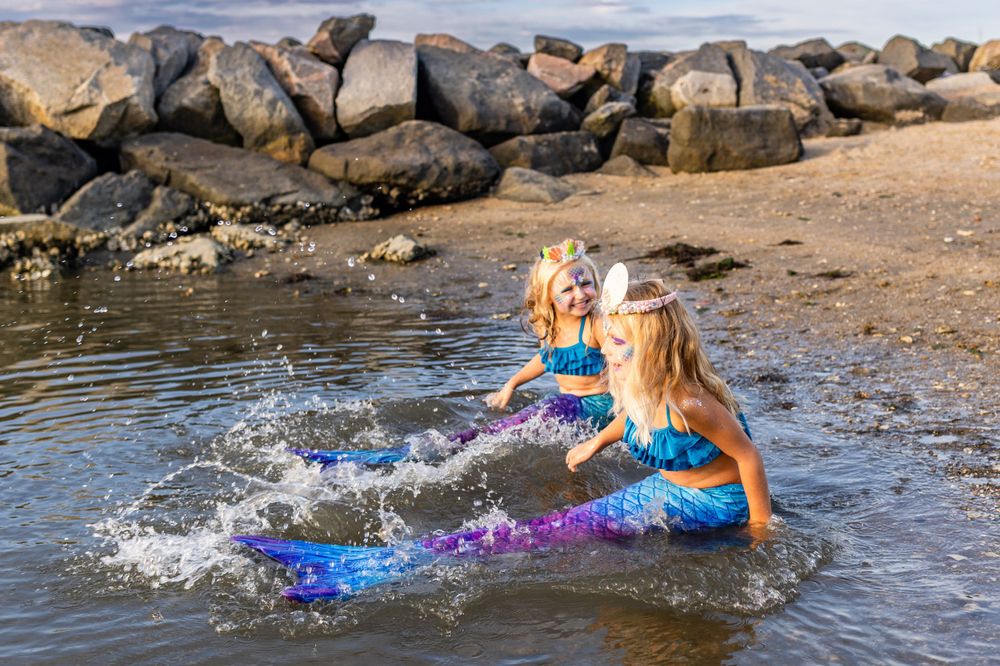 Costumed mermaids splash and play in shallow water near rocky shore.