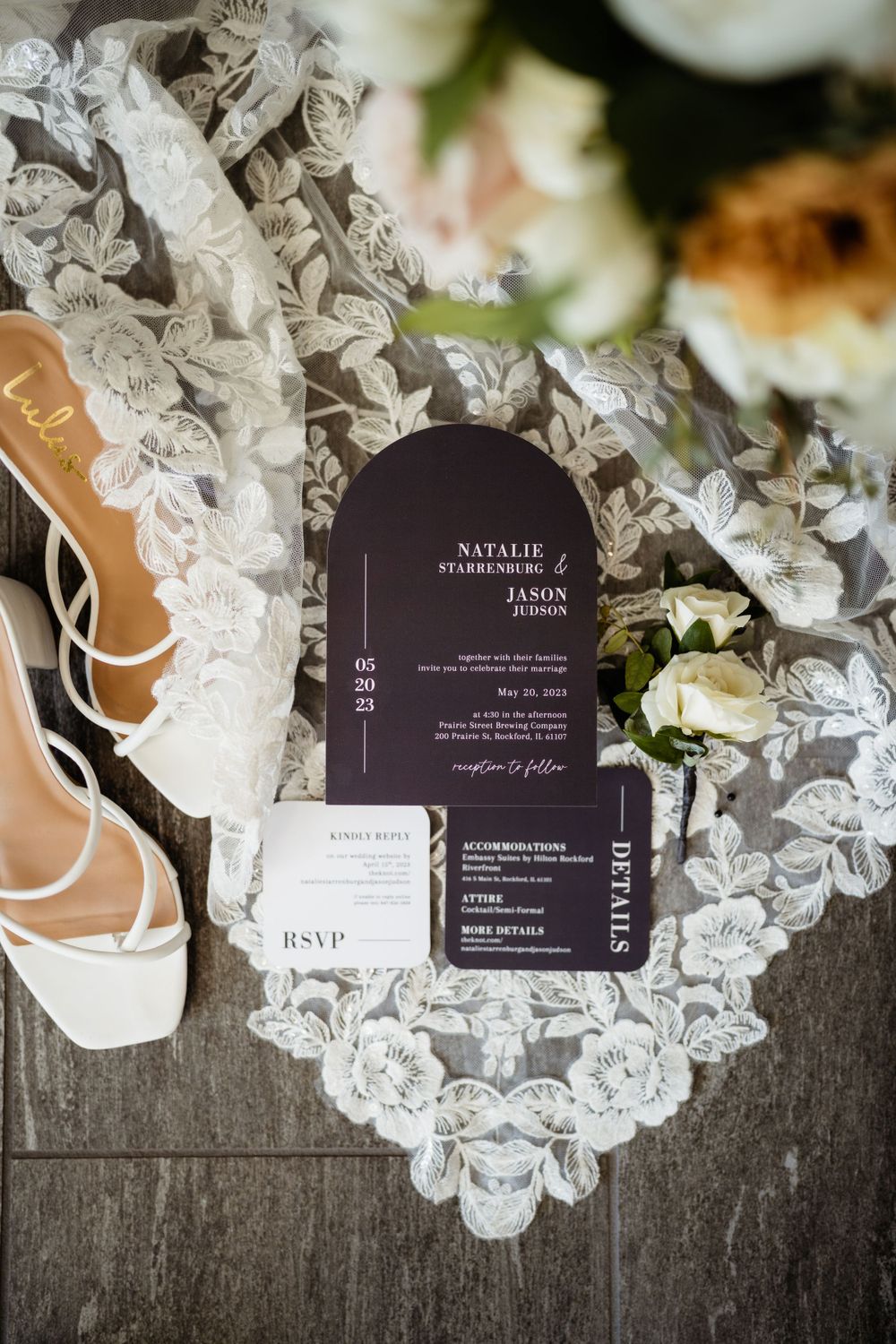 Wedding details including black invitation, delicate lace veil, and white bridal shoes on wooden surface.