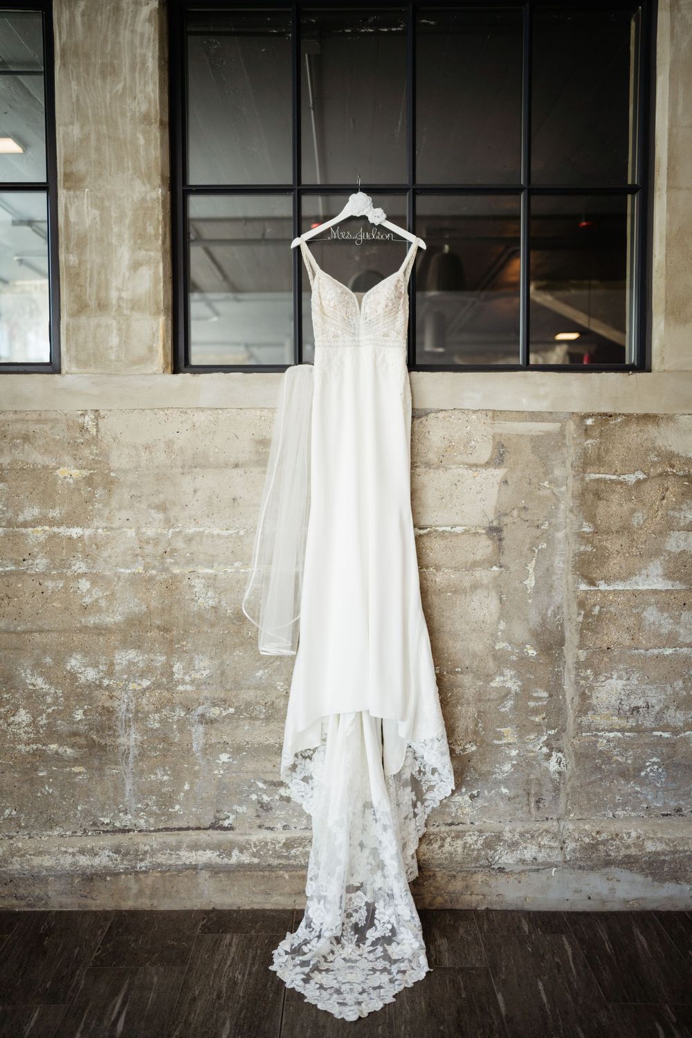 Flowing white wedding gown with lace train suspended against industrial concrete wall.
