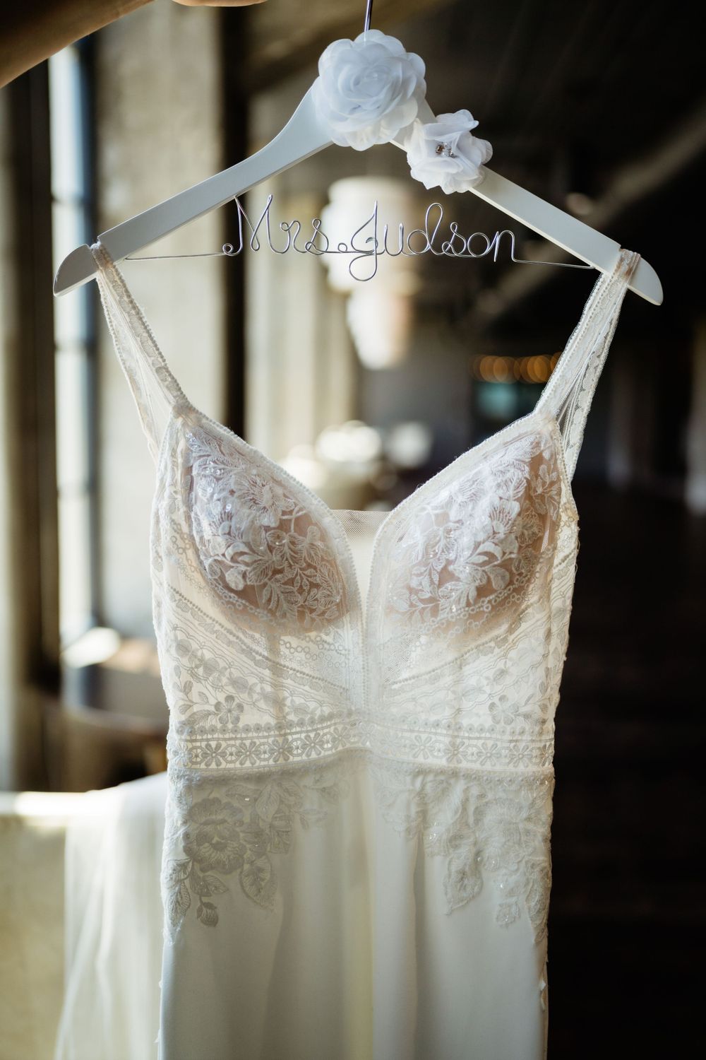 Elegant white lace wedding dress hanging on personalized bridal hanger against soft lighting.