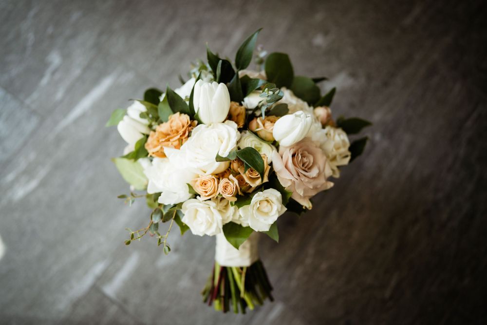 Bridal bouquet featuring cream and peach roses with green foliage on gray background.