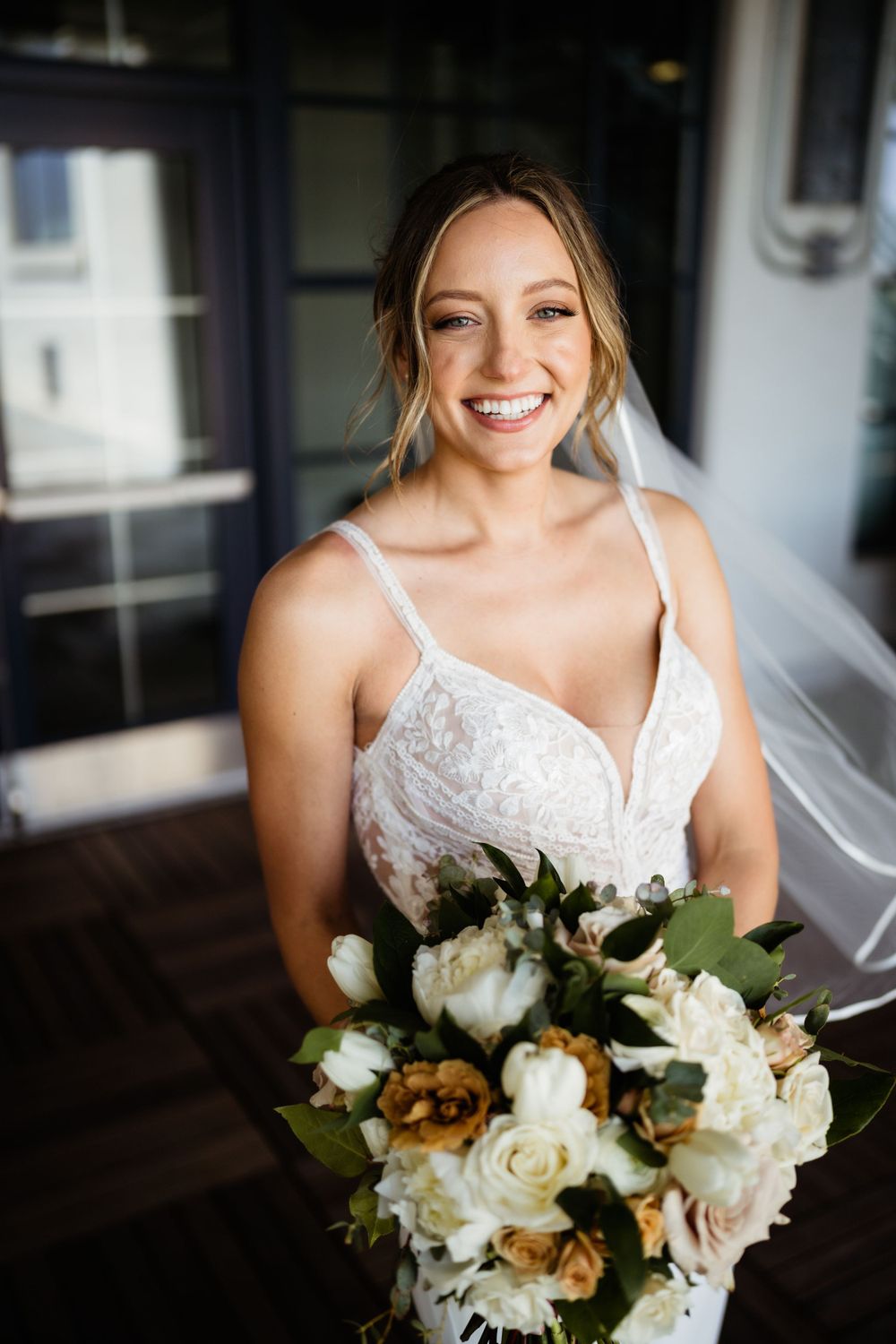 A bride in a white lace wedding dress smiles while holding a beautiful bouquet of white and peach flowers.
