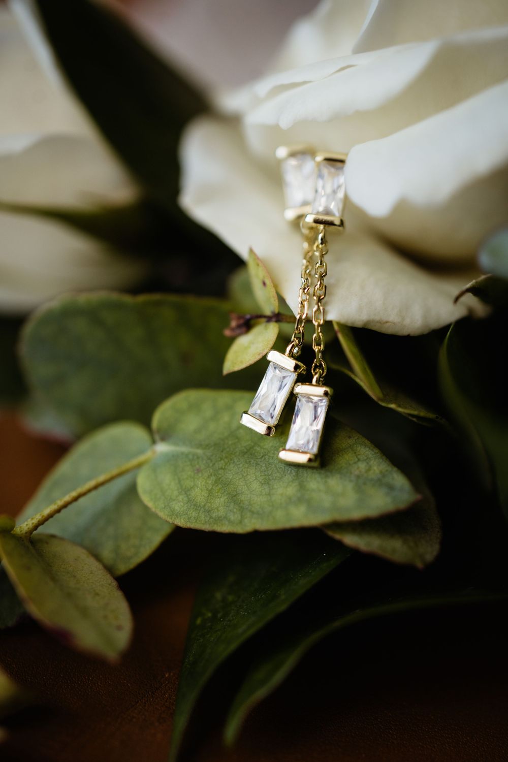 Delicate crystal earrings resting on green eucalyptus leaves with white floral backdrop.