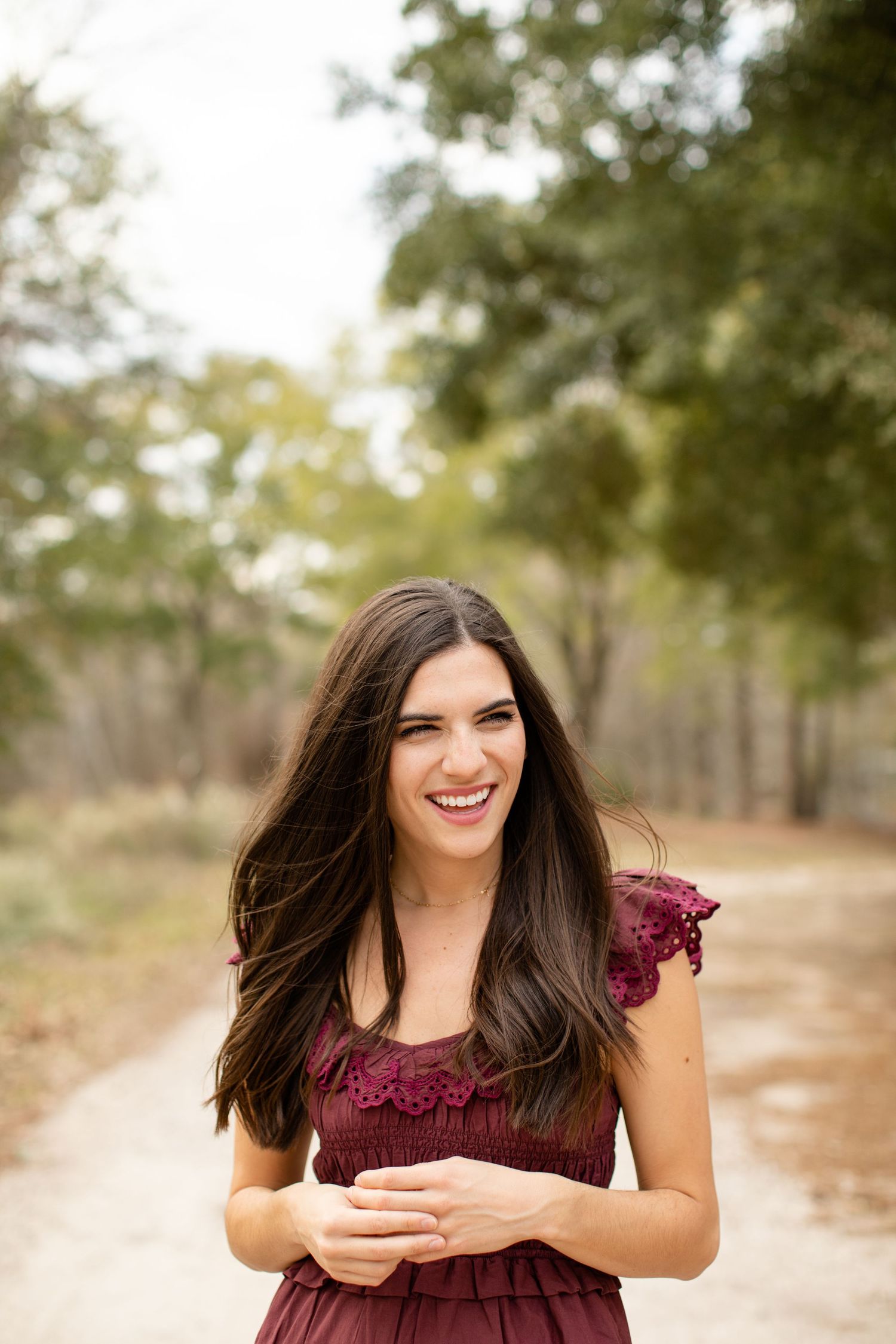 Woman with long dark hair laughs joyfully while wearing a burgundy dress outdoors.